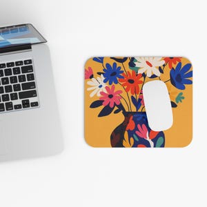 May include: A rectangular mousepad with a floral design featuring a vase filled with colorful flowers. The flowers are in shades of red, blue, pink, and white, set against a yellow background. A white computer mouse is on the right side of the mousepad.