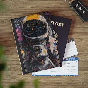 May include: A black passport cover with an astronaut art design, a dark blue passport, and two boarding passes are on a wooden surface. The passport cover features a colorful astronaut illustration with scribbled details. The boarding passes have flight information.
