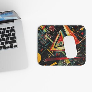 May include: A rectangular mousepad with an abstract geometric design in red, yellow, blue, and white against a dark background. A white computer mouse rests on the pad. A laptop is partially visible in the image.