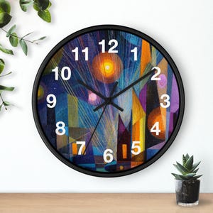 Puede incluir: Un reloj de pared redondo con marco negro. La esfera presenta un diseño abstracto con colores vibrantes, incluyendo azul, naranja, amarillo y morado. Números blancos marcan las horas. Las manecillas del reloj son negras.