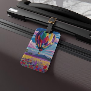 Puede incluir: Una etiqueta de equipaje rectangular que muestra un globo aerostático colorido flotando sobre un campo vibrante de flores rojas y amarillas bajo un cielo nublado. La etiqueta está sujeta al asa de una maleta marrón con una correa negra y una hebilla dorada.