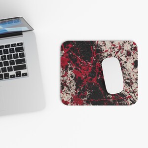 Puede incluir: Una alfombrilla de ratón rectangular con un ratón blanco. La alfombrilla tiene un fondo negro con un diseño de salpicaduras de pintura roja y blanca. Un ordenador portátil plateado está en el fondo.