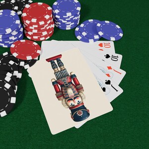 May include: A playing card with a nutcracker design, alongside a hand of cards and stacks of red, blue, and black poker chips on a green surface. The card depicts a nutcracker in red, blue, and white.