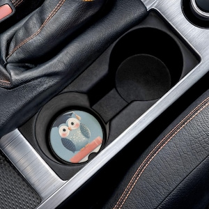 Op de afbeelding: Een auto bekerhouder coaster met een uil ontwerp. De coaster heeft een lichtblauwe achtergrond met een cartoon uil met een afstudeerhoed. De uil heeft grote, ronde ogen en zit op een rode tak.