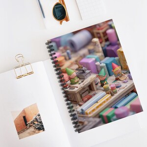 May include: A spiral-bound notebook featuring a whimsical illustration of miniature elves at work in a workshop. The scene includes colorful spools, tools, and figurines, creating a playful and detailed design.