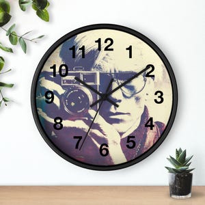 May include: A round wall clock with a black frame featuring a vintage image of a person holding a camera. The clock face displays black numbers and hands against a sepia-toned background. The image has a retro aesthetic.