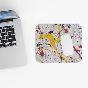 Puede incluir: Una alfombrilla de ratón blanca con un diseño de salpicaduras de pintura abstracta colorida en amarillo, rojo, azul y negro. Un ratón de ordenador blanco está sobre la alfombrilla. Un portátil está en el fondo.