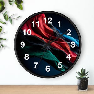 Puede incluir: Un reloj de pared redondo negro con un diseño abstracto colorido. La esfera del reloj presenta un patrón arremolinado de elementos similares a tela roja, azul y verde. Los números blancos marcan las horas sobre un fondo oscuro.