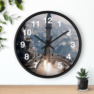 May include: A round wall clock with a black frame featuring a space-themed graphic of a rocket launching from a platform above the clouds. The clock face displays large white numbers from 1 to 12 and has black hour, minute, and second hands.