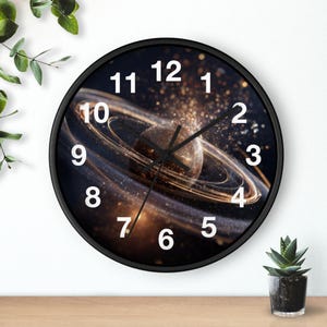 May include: A round wall clock with a black frame and a face featuring a space-themed graphic of a planet with glowing rings and golden particles. The clock has white Arabic numerals from 1 to 12 and black hour, minute, and second hands.