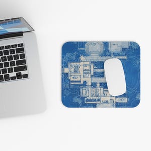 Peut inclure: Un tapis de souris bleu avec un plan architectural détaillé. Une souris d'ordinateur blanche repose sur le tapis, à côté d'un clavier d'ordinateur portable. Le plan montre une disposition de bâtiment avec des détails complexes.