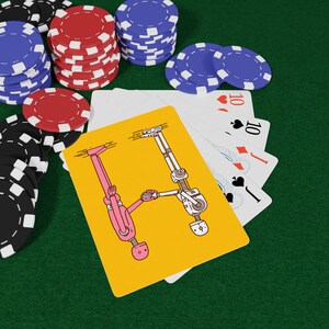 May include: A yellow playing card with a robot illustration, surrounded by poker chips in red, black, and blue, and playing cards on a green surface. The card features a pink robot and a white robot.