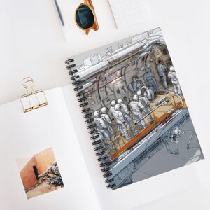 May include: Spiral-bound notebook featuring a detailed illustration of astronauts inside a spacecraft. The artwork depicts figures in white space suits, with intricate mechanical details in shades of gray and orange. The notebook is open on a white surface.