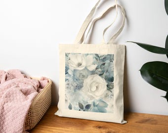 May include: A cream-colored tote bag featuring a floral design with white and blue roses and foliage. The bag has a rectangular print and long handles, perfect for carrying items. The background is a light cream color.