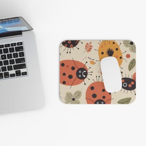 May include: A beige mousepad with a whimsical ladybug pattern. The design features orange and black ladybugs, along with other insect-themed illustrations. A white computer mouse rests on the pad, next to a laptop.