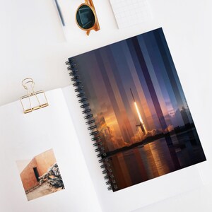 May include: Spiral-bound notebook featuring a vibrant image of a rocket launch against a sunset sky. The cover design incorporates vertical color blocks, adding a modern touch. The notebook is open, revealing blank pages and a gold-colored paper clip.