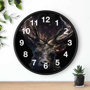 May include: A black round wall clock featuring a detailed image of a stag's head with large antlers. The clock face has white numerals and hands, set against a dark background. The clock is a decorative home decor item.