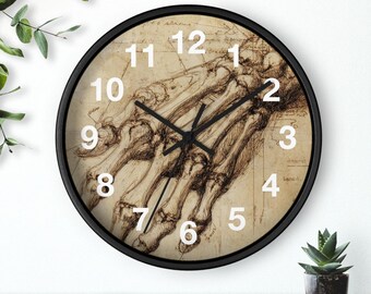 May include: A circular wall clock with a black surround. The clock face displays a sepia-toned anatomical drawing of a hand, with white numerals. Black clock hands show the time.