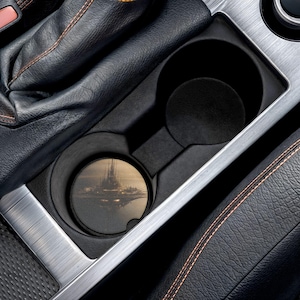 May include: A circular car cup holder coaster featuring a detailed illustration of a futuristic city skyline at dusk. The coaster is placed inside a black plastic vehicle cup holder console next to a black leather gear shift boot with orange stitching.