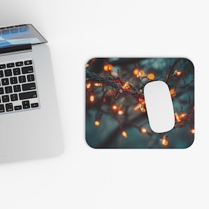 May include: A rectangular mousepad featuring a blurred image of warm, orange fairy lights on a dark background. A white computer mouse is positioned on the right side of the mousepad, next to a laptop.