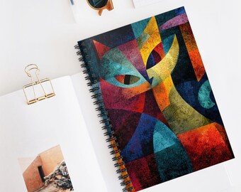 Op de afbeelding: Spiraalgebonden notitieboek met een kleurrijk, geometrisch kattenontwerp. Het kunstwerk maakt gebruik van verschillende kleuren, waaronder blauw, rood, geel en oranje. Het notitieboek ligt open op een wit oppervlak, met een gouden paperclip en zonnebril.