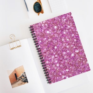 May include: A spiral-bound notebook with a cover covered in pink glitter. The notebook is open on a white surface, with a gold paper clip and sunglasses nearby. The notebook's pages are white.