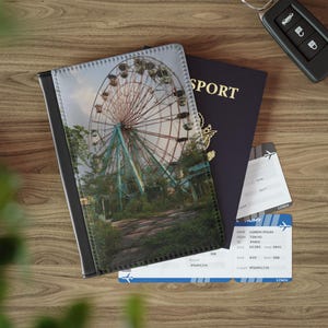 May include: A rectangular passport holder featuring a printed image of a large, weathered Ferris wheel surrounded by overgrown greenery. The holder is positioned next to a dark blue passport, a white boarding pass, and a black car key fob.