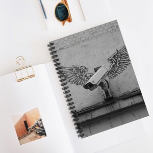 May include: Spiral-bound notebook with a black and white image of a security camera with angel wings graffiti art. The notebook is open, revealing a blank page and a photo. Other items include sunglasses and a gold paper clip.