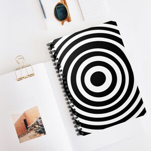Puede incluir: Un cuaderno con espiral con un diseño de círculos concéntricos en blanco y negro. El cuaderno está abierto, revelando una página en blanco. También se ve un clip dorado y gafas de sol.