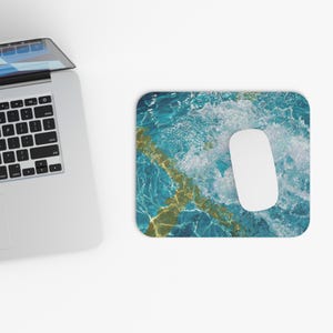 Pode incluir: Um mousepad retangular com um design vibrante de água azul e dourada e um mouse de computador branco. O mousepad é colocado ao lado de um laptop em uma superfície branca. O design da água tem um padrão de ondas.