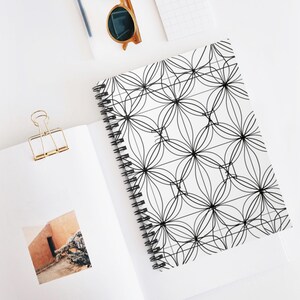 May include: A spiral-bound notebook with a black and white floral pattern. The notebook is open, revealing blank pages. A gold binder clip, sunglasses, and a pen are also visible on the white surface.