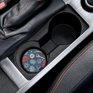 May include: A car coaster with a navy blue background featuring a pattern of gears, wrenches, and red hard hats. The coaster is placed inside a car's cup holder, surrounded by black leather and silver trim.