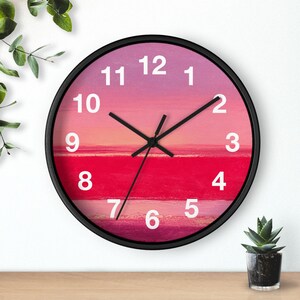 May include: A round wall clock with a black frame. The clock face features a gradient of pink and red hues, resembling a sunset over the ocean. White numerals mark the hours, and black hands indicate the time.