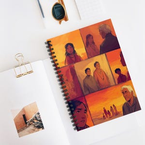 May include: Spiral-bound notebook with a comic-style illustration in warm orange and red hues. The artwork shows various figures in a series of panels. The notebook is open on a white surface, with a gold paper clip and sunglasses nearby.