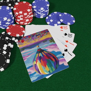 Può includere: Una carta da gioco con un'illustrazione colorata di una mongolfiera, disposta a ventaglio con altre carte che mostrano il dieci di cuori, dieci di picche, fante di quadri e fante di picche, circondata da pile di fiches rosse, blu e nere.