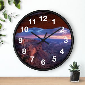 Könnte beinhalten: Eine runde Wanduhr mit schwarzem Rahmen und einem Zifferblatt, das eine Landschaftsaufnahme eines Canyons bei Sonnenuntergang zeigt. Die Uhr hat weiße arabische Ziffern von 1 bis 12 sowie schwarze Zeiger für Stunden, Minuten und Sekunden.