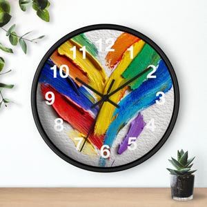 Może przedstawiać: Okrągły zegar ścienny z czarną ramą. Tarcza zegara przedstawia wzór w kształcie serca z kolorowymi pociągnięciami pędzla w odcieniach czerwieni, pomarańczy, żółci, zieleni, niebieskiego i fioletu. Godziny oznaczone są białymi cyframi.