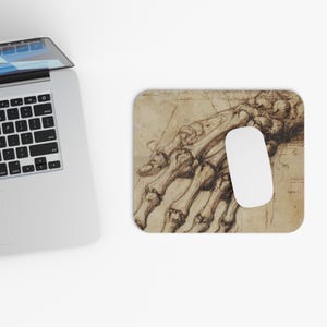 Può includere: Un tappetino per mouse rettangolare con un disegno anatomico dettagliato di una mano in tonalità seppia. Un mouse per computer bianco è appoggiato sul tappetino, accanto a un laptop.