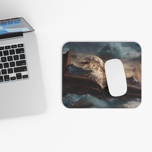 May include: A rectangular mouse mat with an open book design depicting a stormy sea and a city illustration. A white computer mouse sits on the mat, beside a laptop.