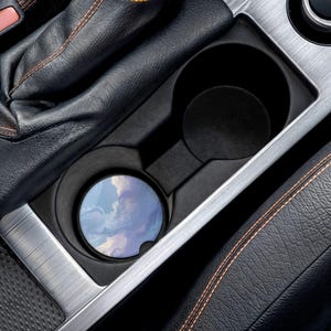 May include: A car cup holder with a round coaster featuring a blue and white cloud design. The coaster is placed inside the cup holder, which is surrounded by black leather and silver trim.