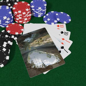 May include: A hand of playing cards featuring a landscape image of a rocky canyon with a reflection of trees and sky in a pool of water, fanned out alongside stacks of red, blue, and black poker chips on a green felt surface.