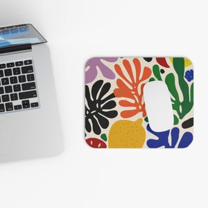 May include: A rectangular mousepad with a colorful abstract design featuring various shapes in orange, green, purple, yellow, and black. A white computer mouse sits in the center of the mousepad. A laptop is partially visible in the image.