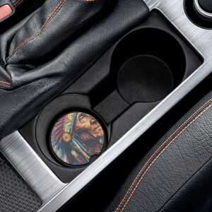 May include: A car cup holder with a colorful round coaster featuring a Native American chief portrait. The coaster is placed inside the cup holder, which is part of a black interior with leather and silver accents.