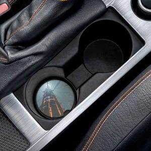 May include: A car cup holder with a coaster featuring a cityscape image. The coaster is round and fits snugly in the cup holder. The interior of the car is black leather with orange stitching and a silver trim.