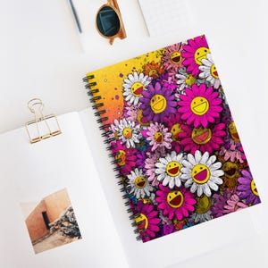 Può includere: Un quaderno a spirale con un vivace design floreale. La copertina presenta un collage di fiori sorridenti nei toni del rosa, viola, bianco e giallo su uno sfondo sfumato. Il quaderno è aperto, rivelando pagine bianche e una graffetta dorata.