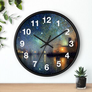 Op de afbeelding: Een ronde wandklok met een zwart frame. De klok heeft een wijzerplaat met een sterrennachtscène met een waterkant. Witte cijfers markeren de uren en zwarte wijzers geven de tijd aan.