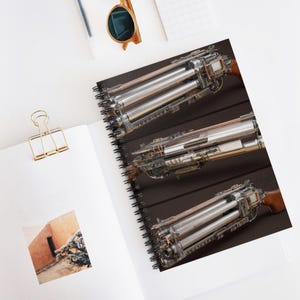 Puede incluir: Cuaderno de espiral con una ilustración detallada de un dispositivo mecánico con múltiples cañones. El dispositivo tiene un acabado metálico con detalles de madera. El cuaderno está abierto sobre una superficie blanca, con un clip dorado y gafas de sol visibles.