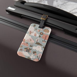 May include: A rectangular luggage tag with a light blue background featuring a repeating pattern of pink and brown stylized trees, clouds, and small white rabbit figures. The tag is attached to a dark brown suitcase handle by a black strap with a gold buckle.