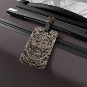 May include: A rectangular luggage tag featuring a detailed, monochromatic stone-like relief pattern with intricate floral and scrollwork carvings. The tag is attached to a dark brown suitcase handle by a black strap with a gold-toned metal buckle.