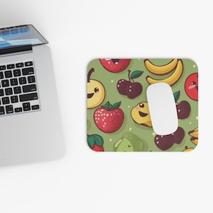 Pode incluir: Um tapete de rato retangular verde com um padrão de frutas de desenho animado sorrindo, incluindo morangos, cerejas, bananas e peras. Um rato de computador branco repousa sobre o tapete, ao lado de um laptop.
